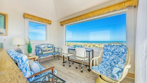 Une pièce dotée d’une grande fenêtre donnant sur une plage, d’un plafond en bois, d’une table avec des chaises et d’un canapé agrémenté d’un coussin sur le thème de la plage.