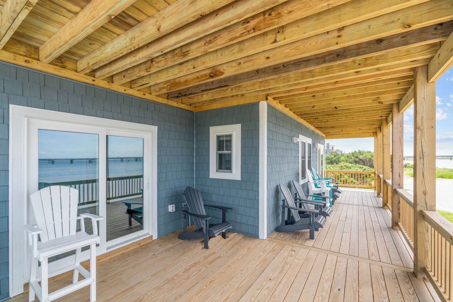 A covered deck with wooden flooring, two white Adirondack chairs, and a view of the ocean.