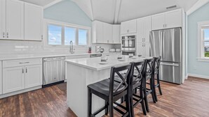 Una cocina moderna con armarios blancos, una isla central con taburetes de bar, electrodomésticos de acero inoxidable y vistas al cielo a través de amplios ventanales.