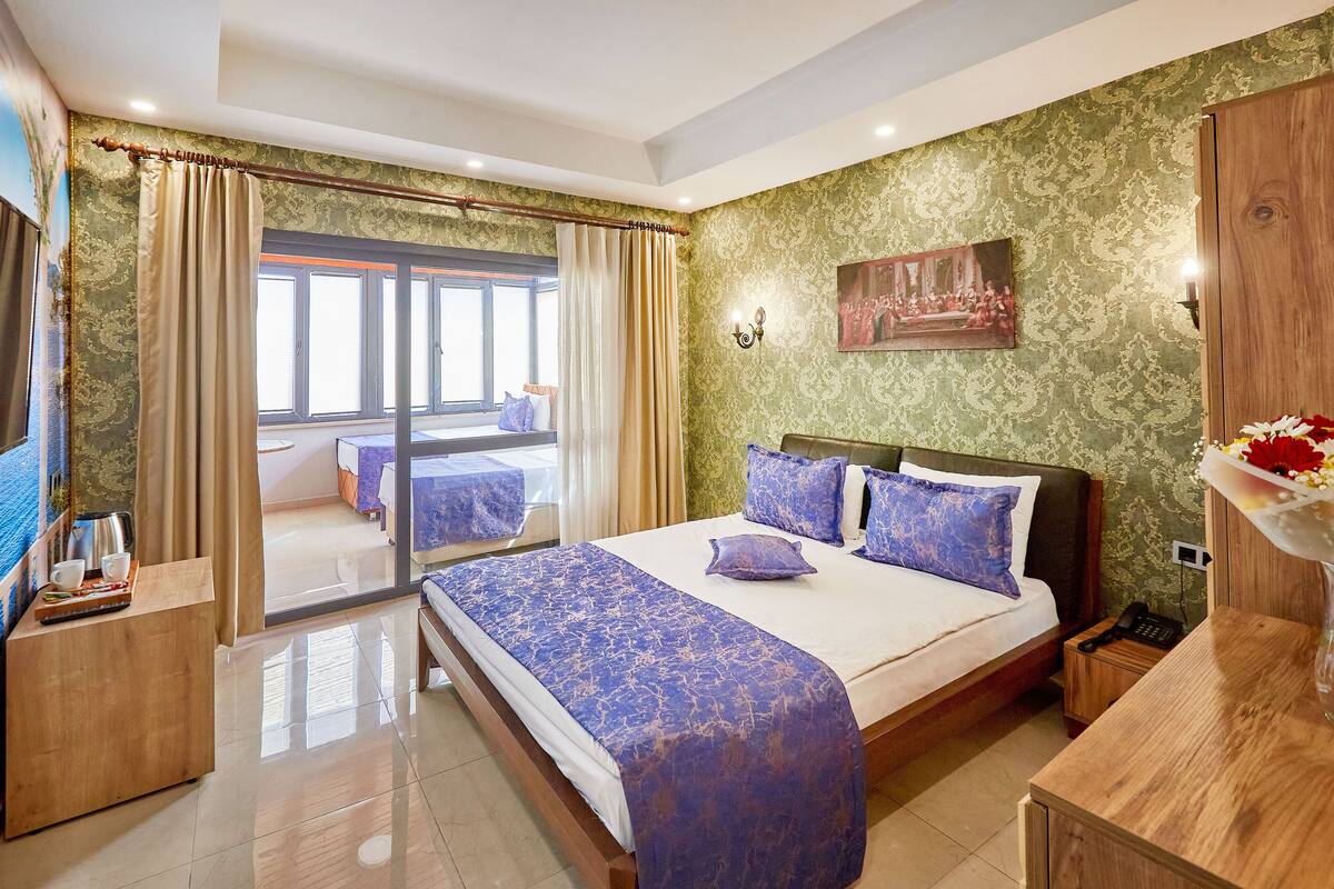 Habitación de hotel con una cama grande, una mesita de noche de madera, un escritorio y vistas al balcón.