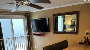A dining area with a wooden table and chairs, a ceiling fan, a flat-screen TV mounted on the wall, and a balcony with a railing.