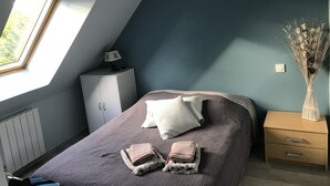 Une chambre avec un plafond en pente, un lit avec des oreillers et des serviettes pliées, une table de chevet avec une lampe, un radiateur et une fenêtre donnant sur un paysage verdoyant.