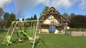 Une maison en bois avec un balcon, une balançoire et un toboggan dans une cour verdoyante.