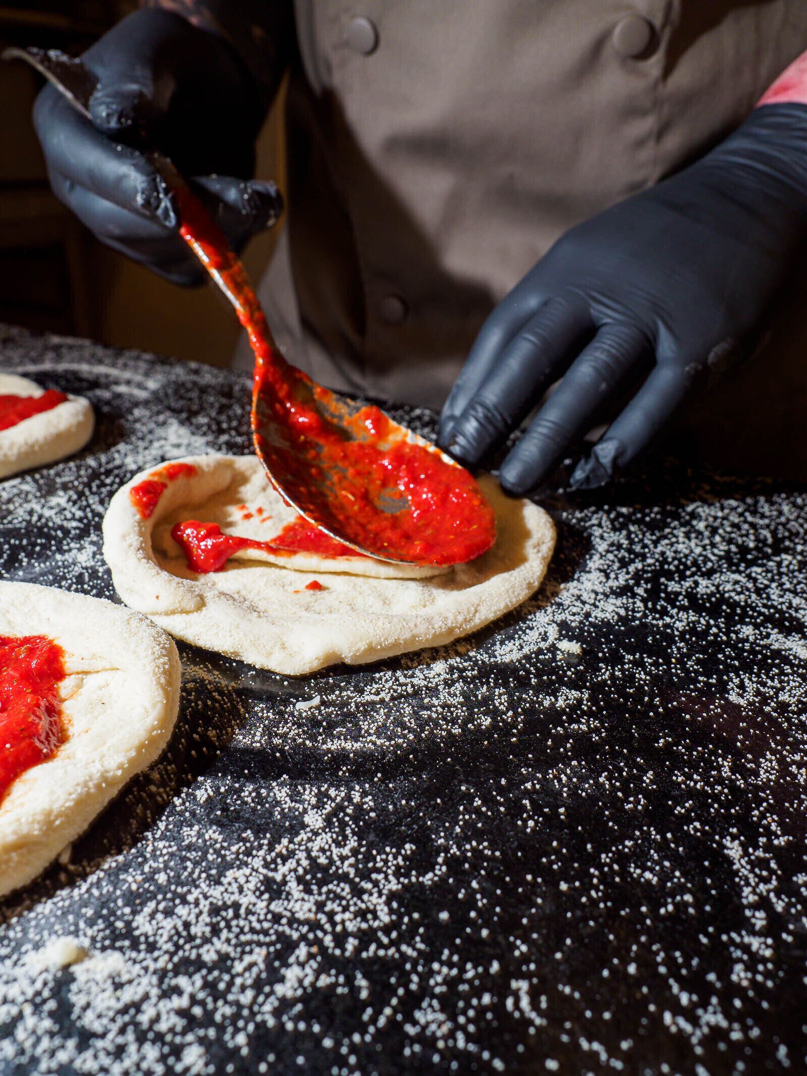 A person in a black chef's coat and gloves preparing food on a black surface with powdered sugar.