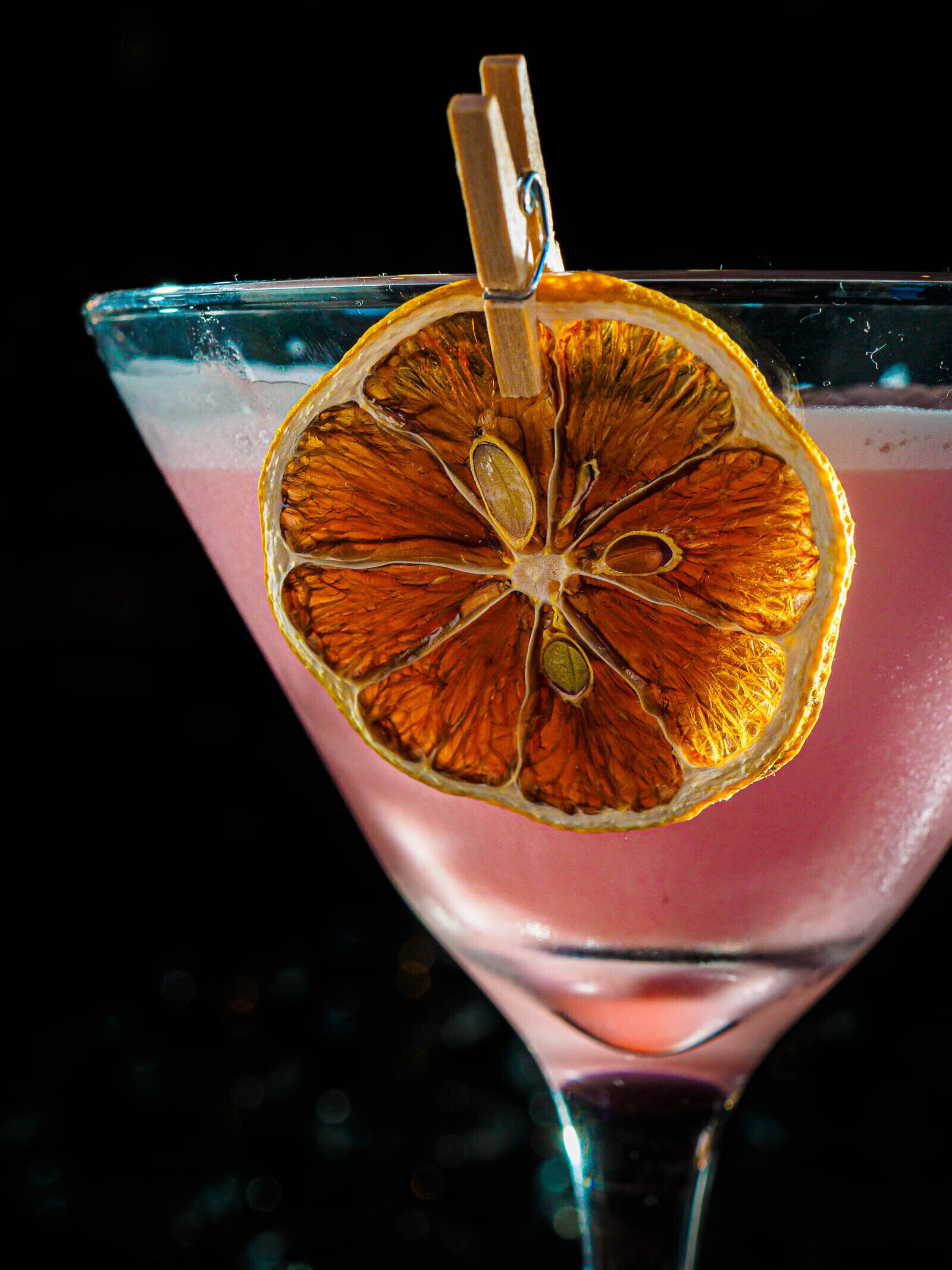 Un verre à martini, garni d’une tranche d’orange séchée et sur lequel est posé un trombone à linge, le tout sur un fond sombre.