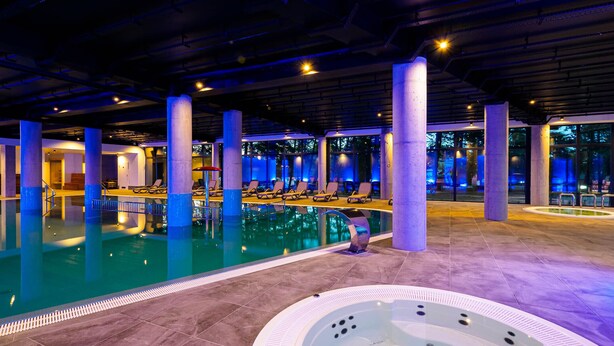 An indoor swimming pool with lounge chairs and a hot tub, surrounded by large pillars and glass walls. - Resort Król Plaza Spa & Wellness (Postomino)