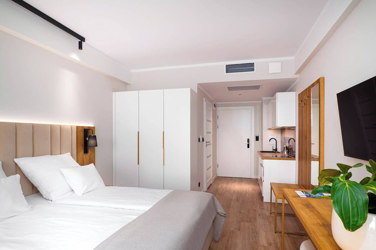 A modern hotel room with a bed, wardrobe, desk, and kitchenette.