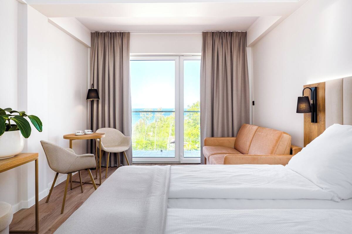 A hotel room with a bed, a desk, a chair, a table, a lamp, and a view of the outdoors.