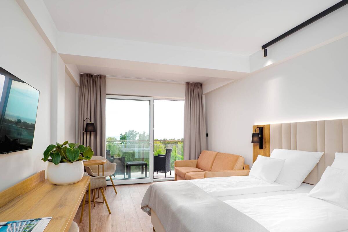 A hotel room with a bed, a desk with a plant, a chair, a sofa, and a balcony with a view.
