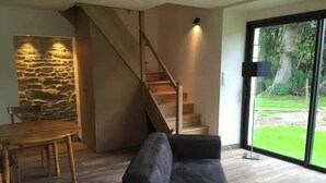 A modern living room with a wooden dining table, a dark sofa, a staircase, and a large glass door leading to a garden. - Petite Maison - Piscine & Vue mer Grange Ouest (Locoal-Mendon)
