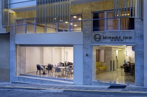 Ένα διώροφο κτίριο με γυάλινη πρόσοψη, ένα ορατό τμήμα εστιατορίου με τραπέζια και καρέκλες και μια επιγραφή που γράφει «Hypnos Inn».