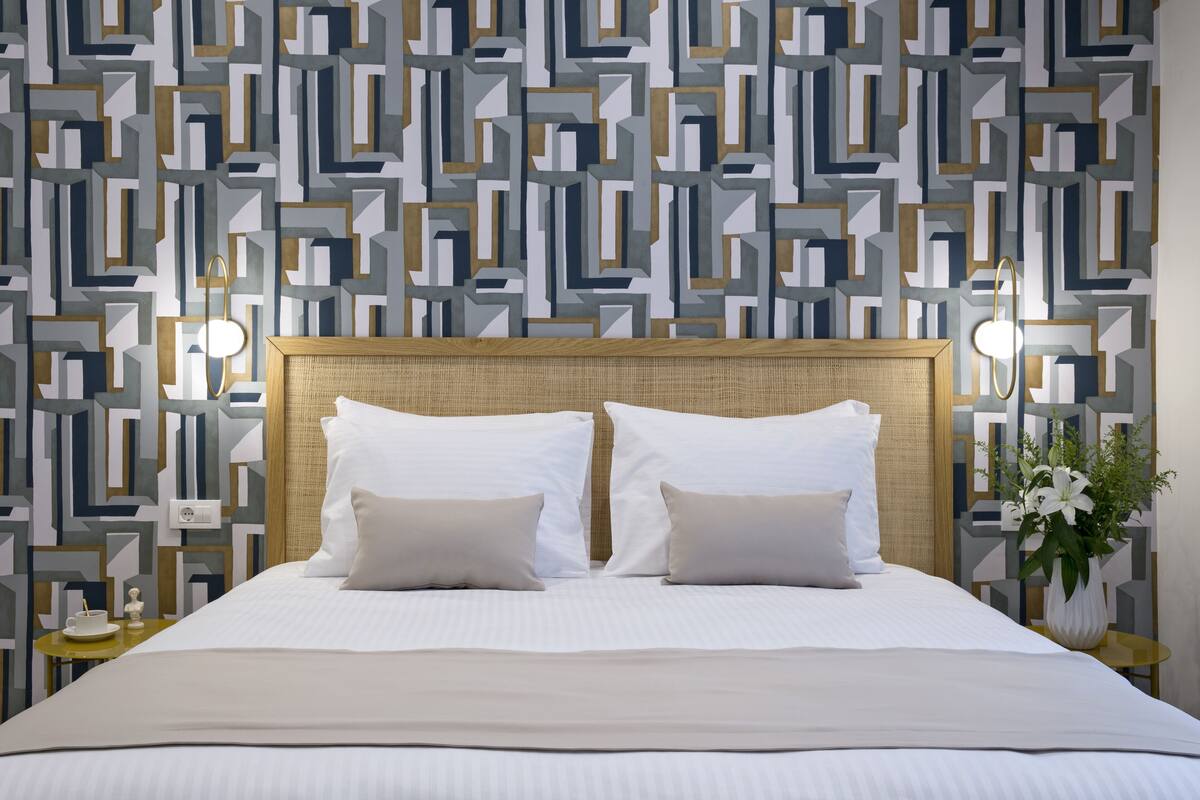 A neatly made bed with a geometric patterned wallpaper, a wicker headboard, and a small table with a vase of flowers.