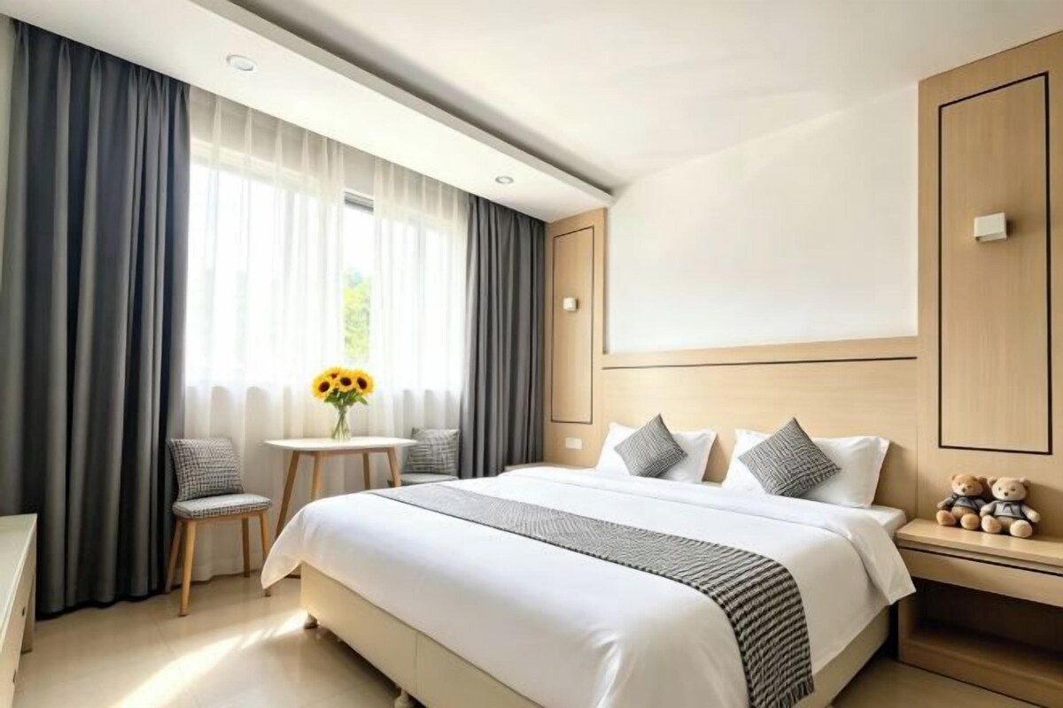 A hotel room with a large bed, a small table with a vase, and a nightstand with a teddy bear.