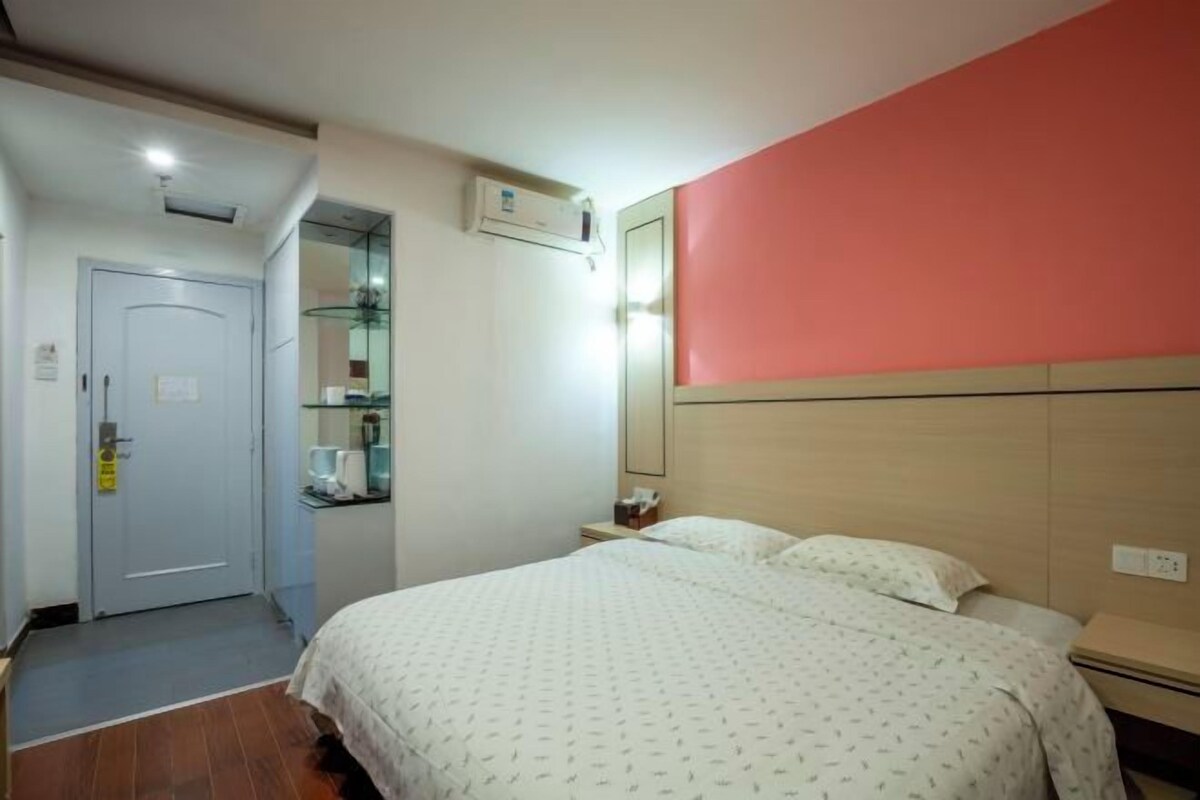 A hotel room with a bed, bedside table, and a small desk with a chair. There is a door to the left and a mirror on the wall.