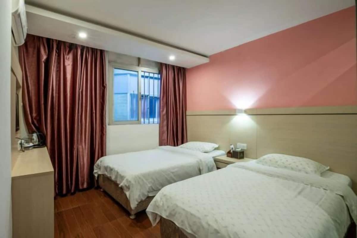 A hotel room with two beds, a window with curtains, a nightstand, and a wall-mounted light.