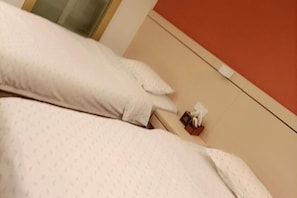 A bed with white bedding and pillows, a bedside table with a tissue box and a small item on it, and a red wall in the background. - Longxiang Hotel (Zhuhai)