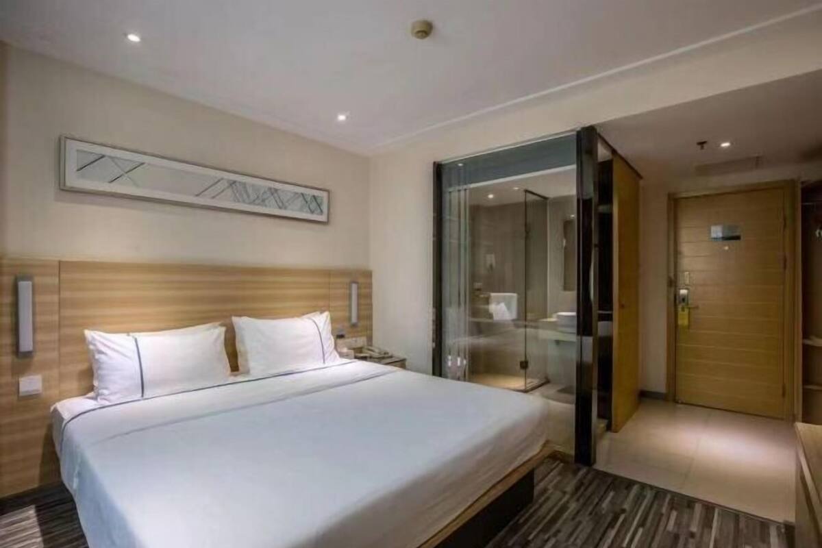 A hotel room with a large bed, a glass partition, a wooden headboard, and a modern light fixture.