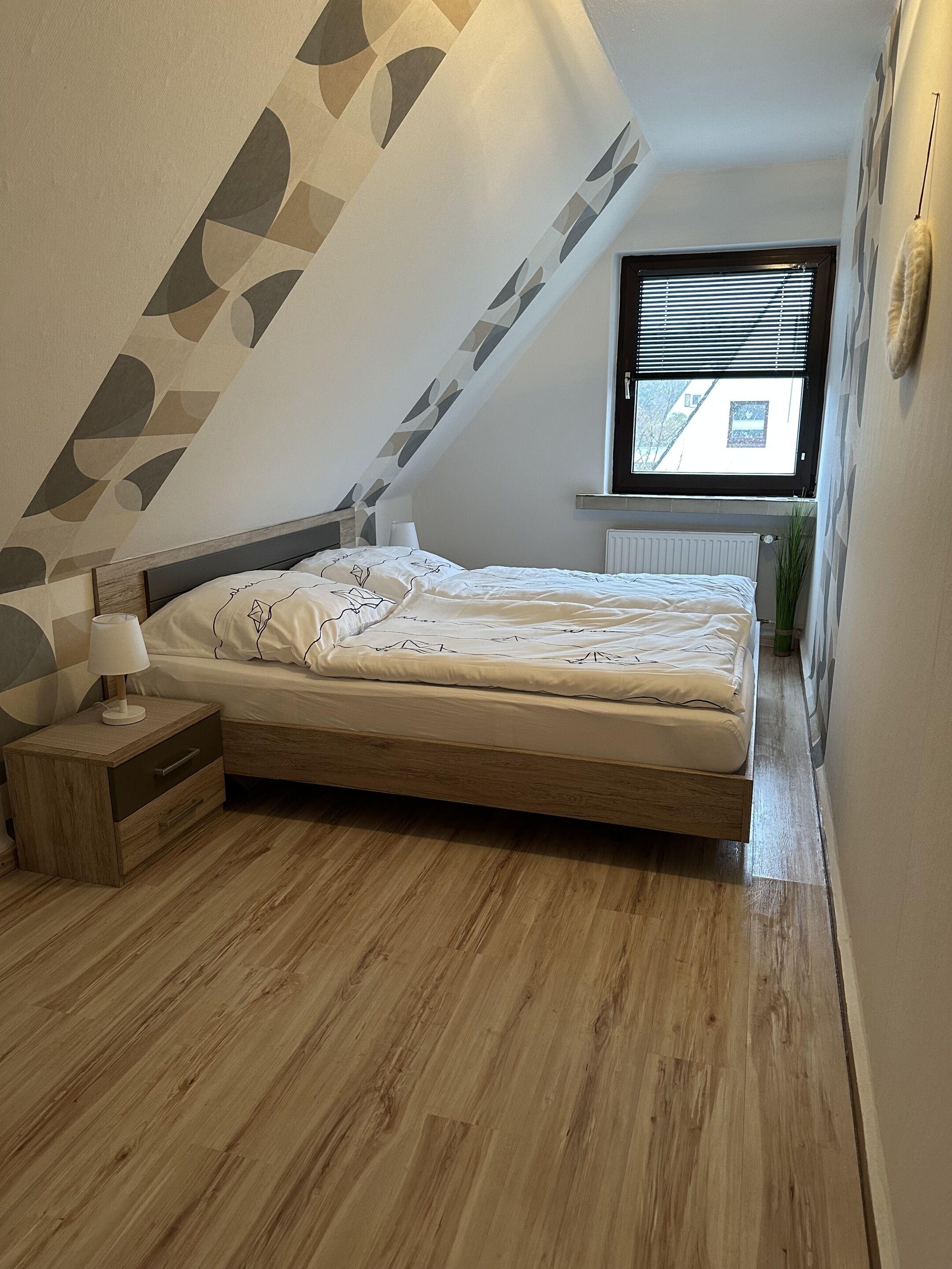 A bedroom with a sloped ceiling, a bed with a white mattress and patterned bedding, a bedside table with a lamp, and a window with blinds.