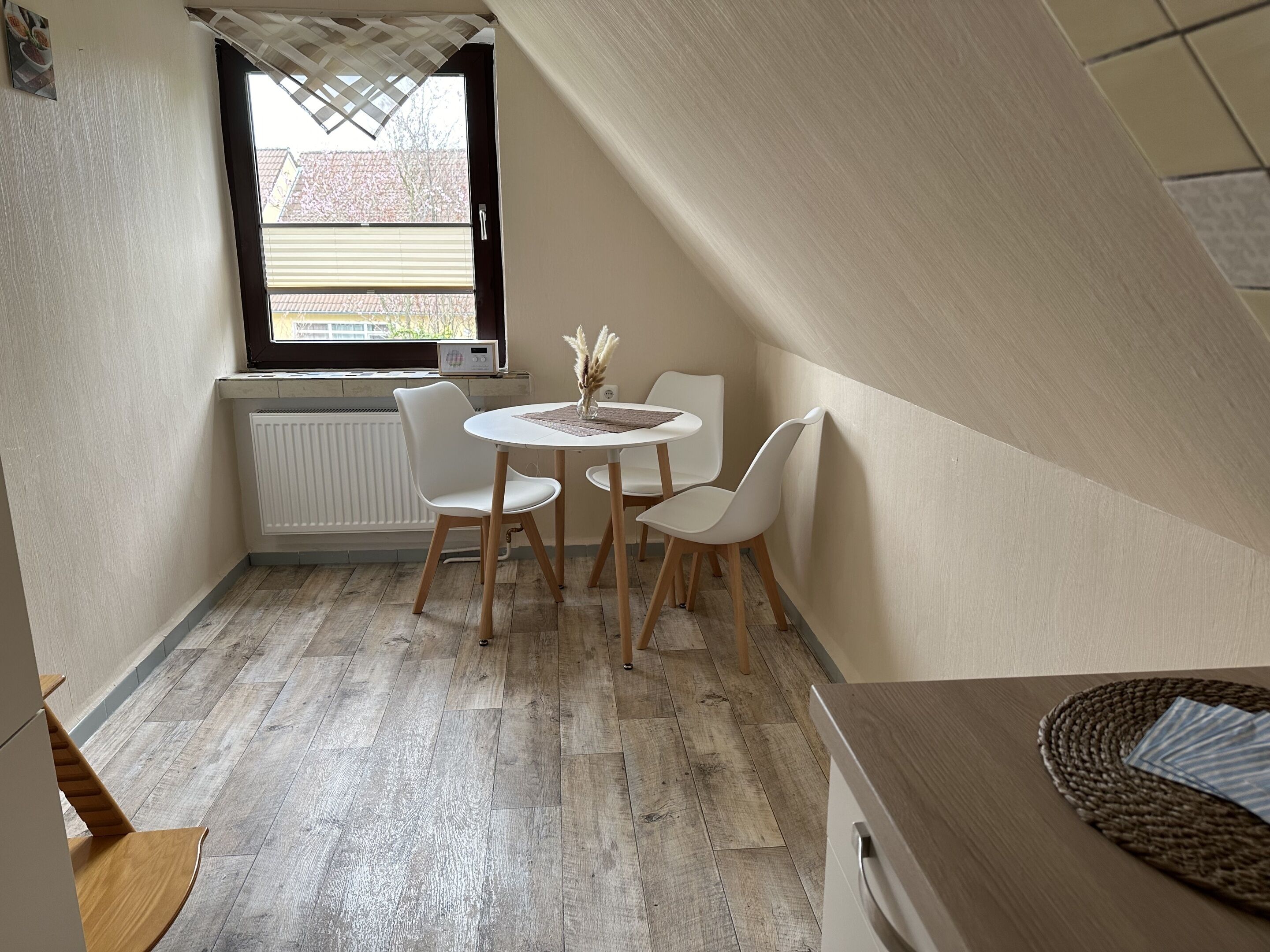 A small, cozy room with a round table and two chairs, a radiator, a window with blinds, and a sloped ceiling.