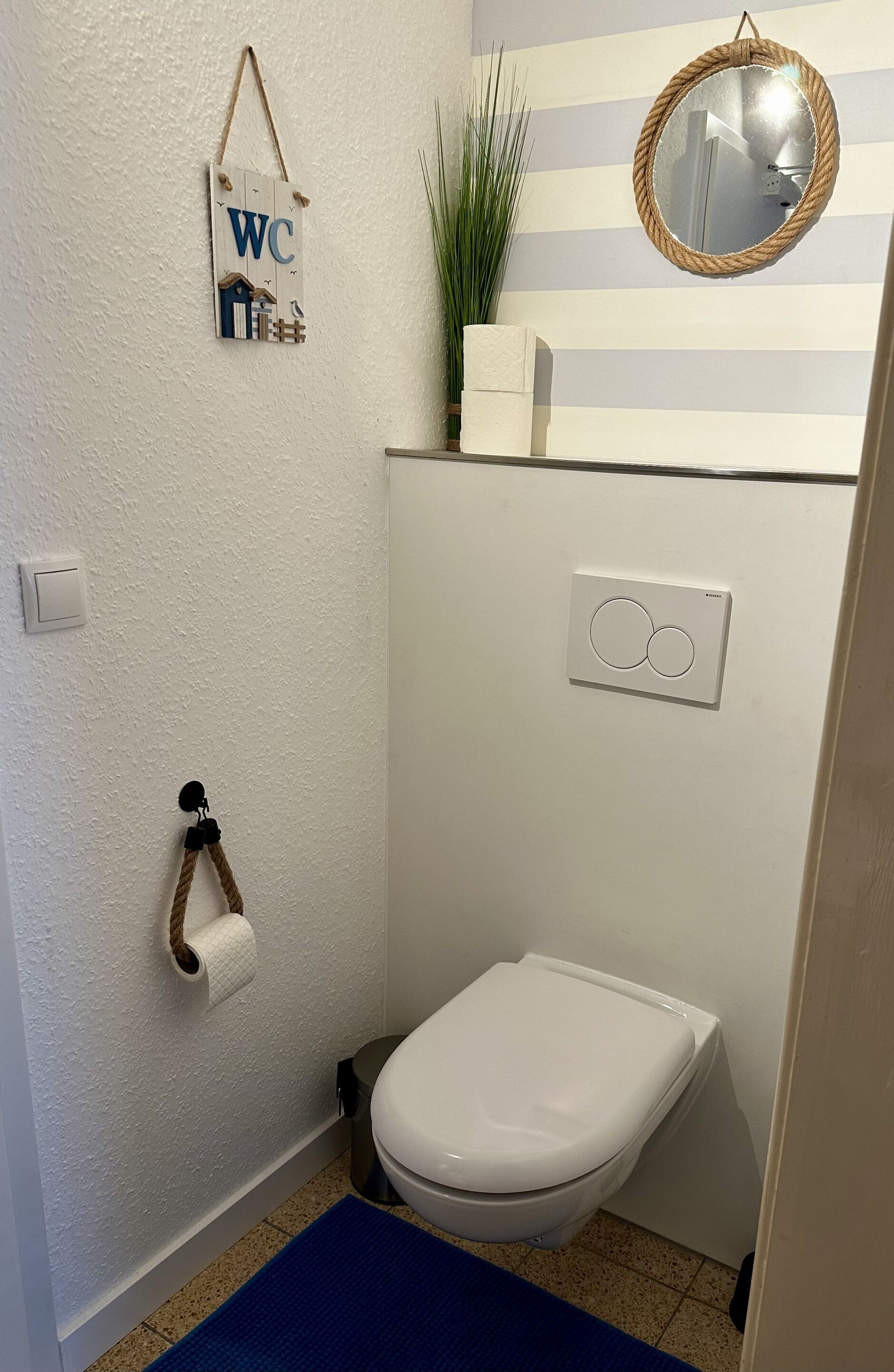A small bathroom with a toilet, a hanging toilet paper holder, a wall-mounted mirror, and a 'WC' sign.