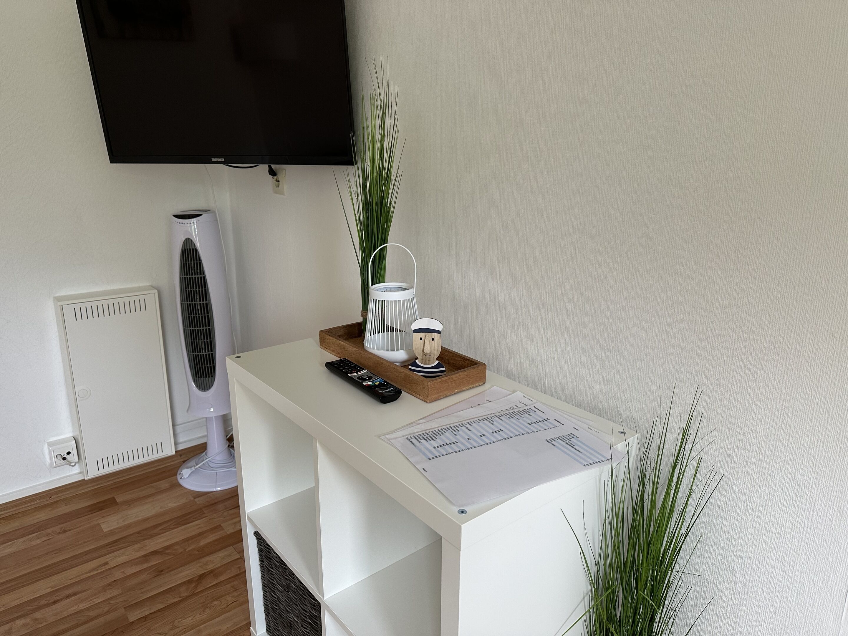 A modern living room with a white entertainment unit, a flat-screen TV mounted on the wall, a standing fan, and a small wooden table with decorative items.
