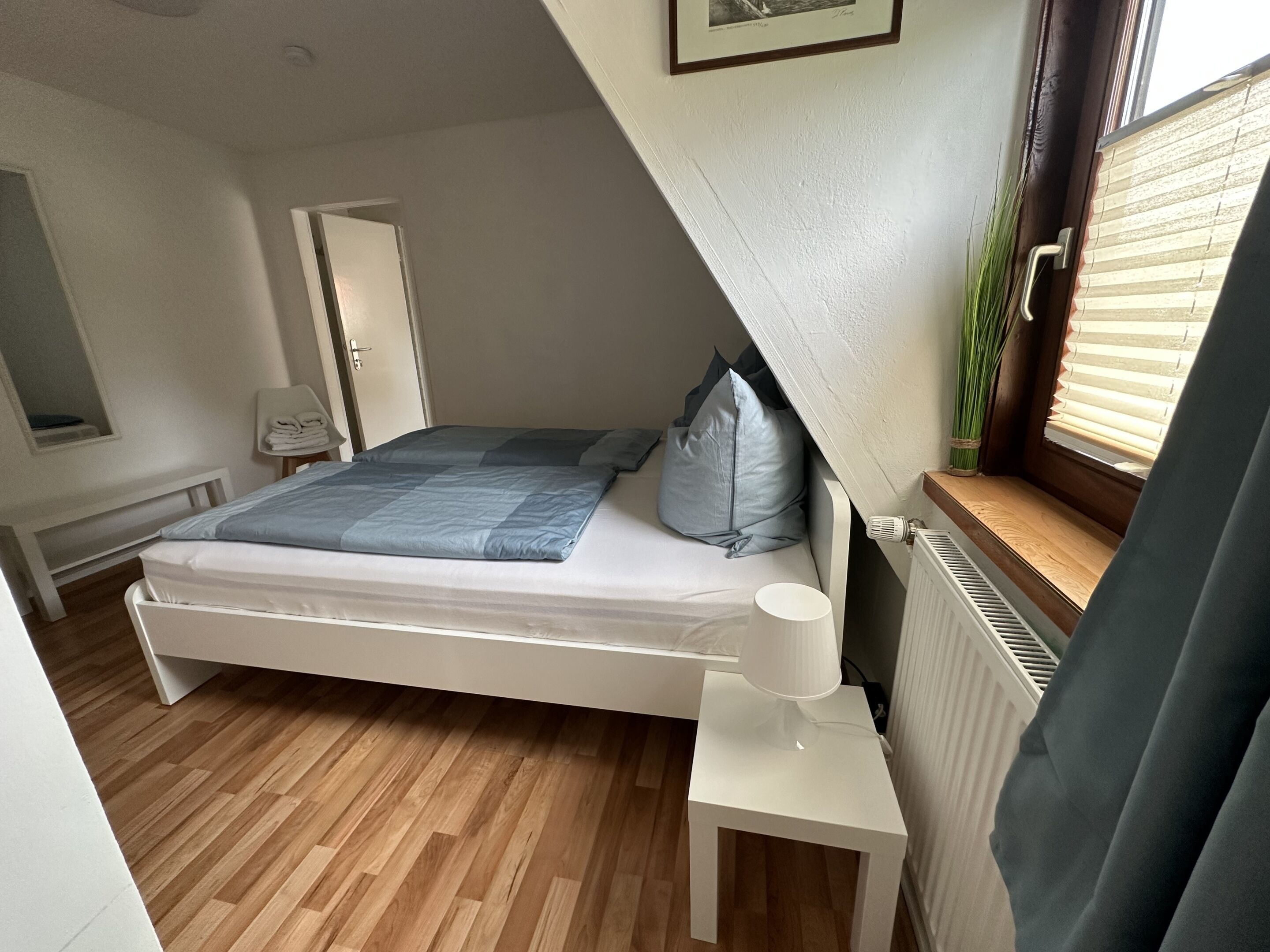 A bedroom with a bed, a bedside table, a radiator, a window with blinds, and a door.