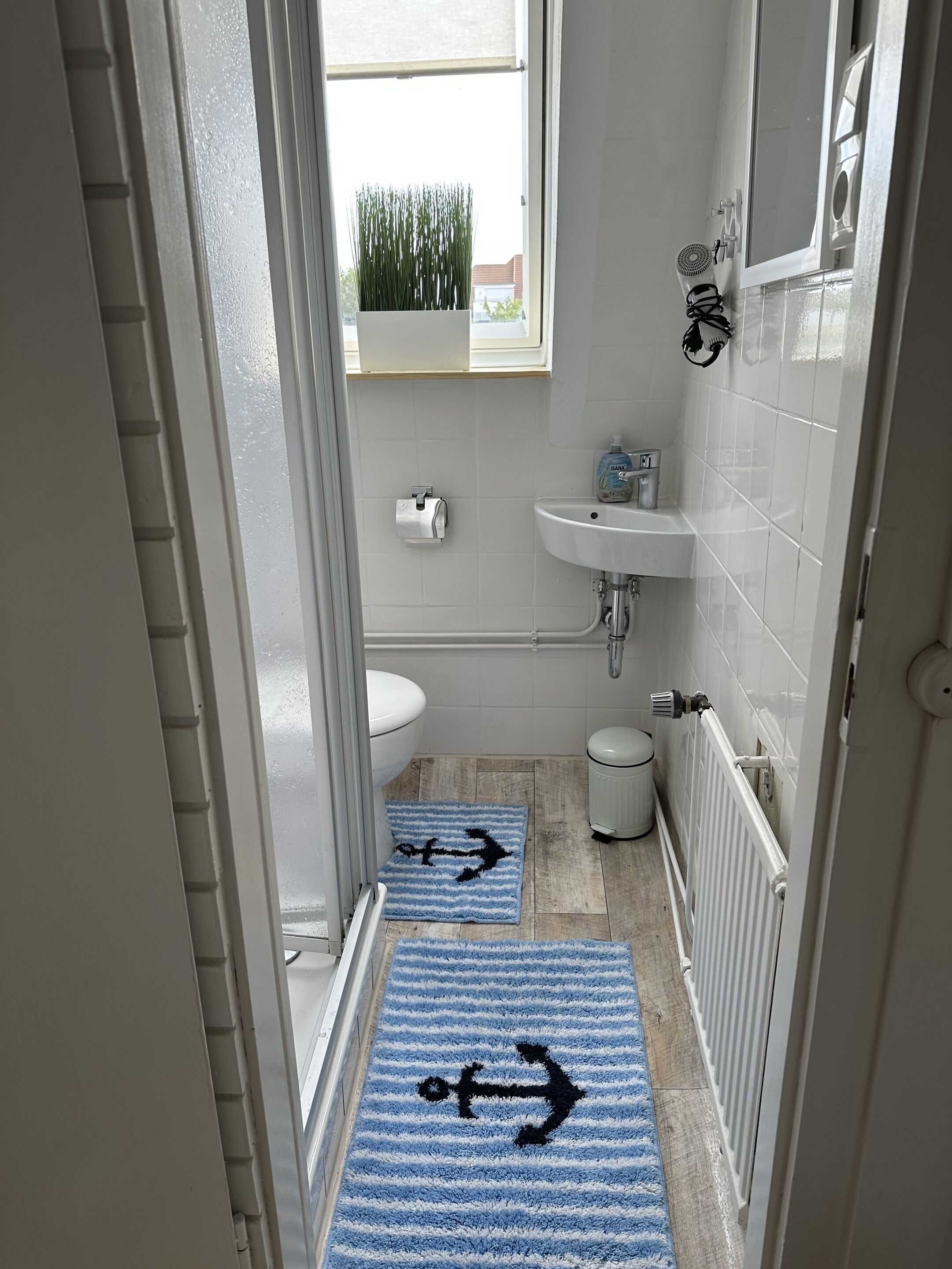 Un baño con inodoro, lavabo y radiador. Hay felpudos azules con diseño de anclas en el suelo.
