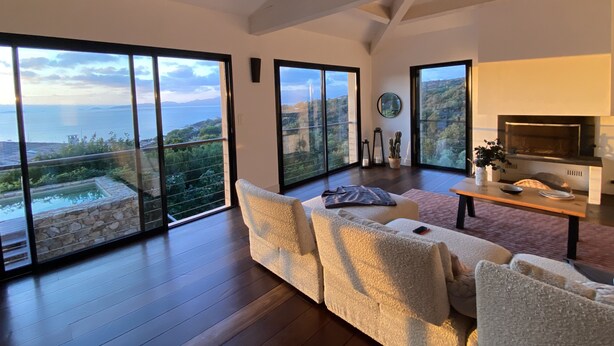Un salón espacioso con un sofá grande, una mesa de centro y una chimenea. La habitación tiene grandes puertas correderas de vidrio que dan a un balcón con vistas al mar.
