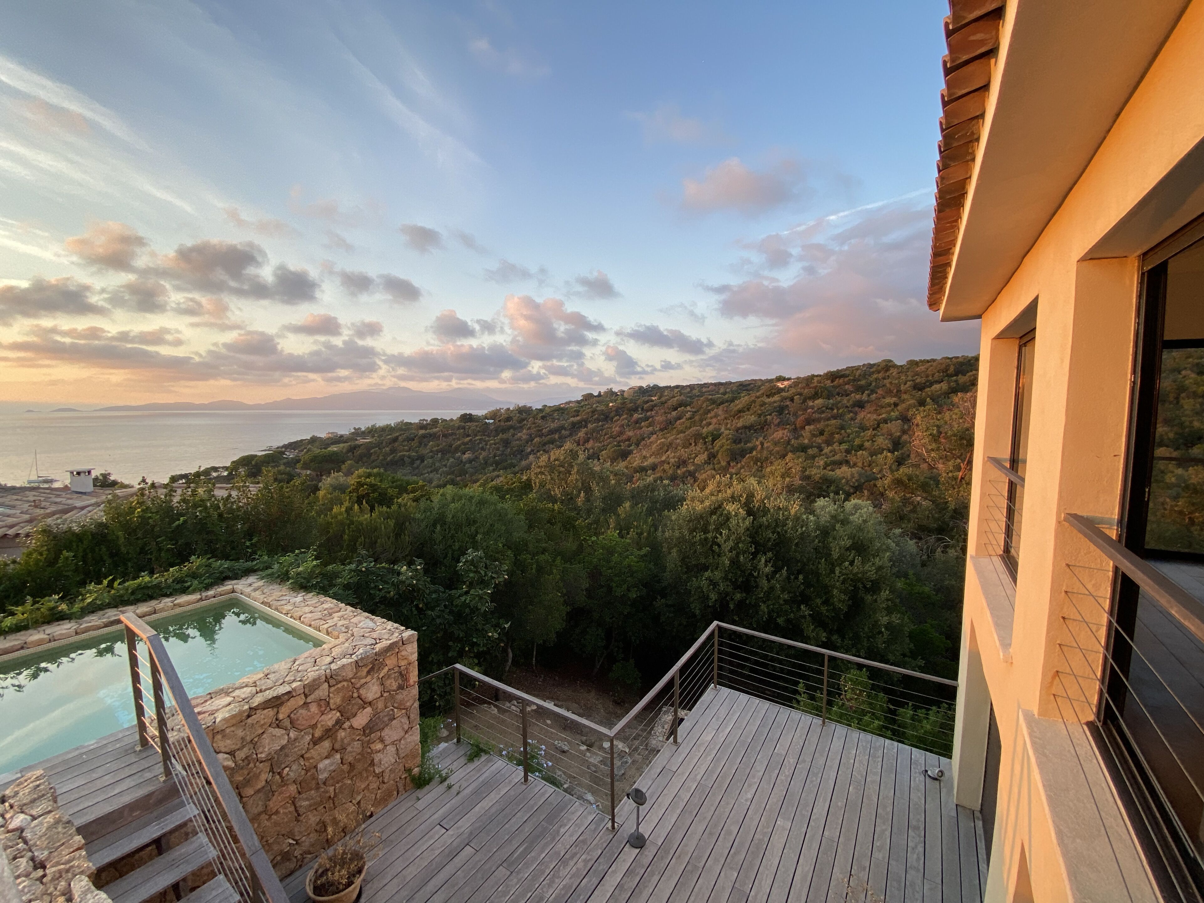 A wooden deck with a stone pool, overlooking a hillside with dense vegetation and a view of the sea.
