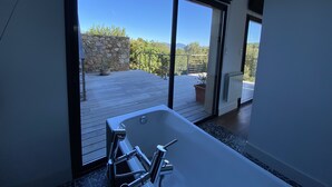 Un baño moderno con una bañera grande, una pared de piedra y vistas a una terraza de madera y vegetación.