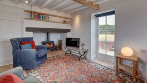 A living room with a blue armchair, a fireplace, a television, and a large window. - Detached 1-bed cottage overlooking the salt marshes (Salthouse)