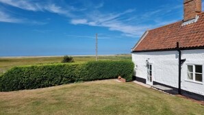 A white house with a red-tiled roof, surrounded by a well-maintained lawn and a hedge. - Detached 1-bed cottage overlooking the salt marshes (Salthouse)