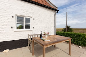 A wooden table with a bottle and flowers on a white wall with a window. - Detached 1-bed cottage overlooking the salt marshes (Salthouse)