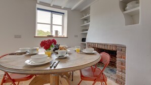 A dining area with a round wooden table, red chairs, white plates, cups, and a vase with red flowers. - Detached 1-bed cottage overlooking the salt marshes (Salthouse)