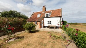 A two-story house with a red-tiled roof, a brick chimney, and a white exterior. There is a small garden with flowering plants and a bench. - Detached 1-bed cottage overlooking the salt marshes (Salthouse)
