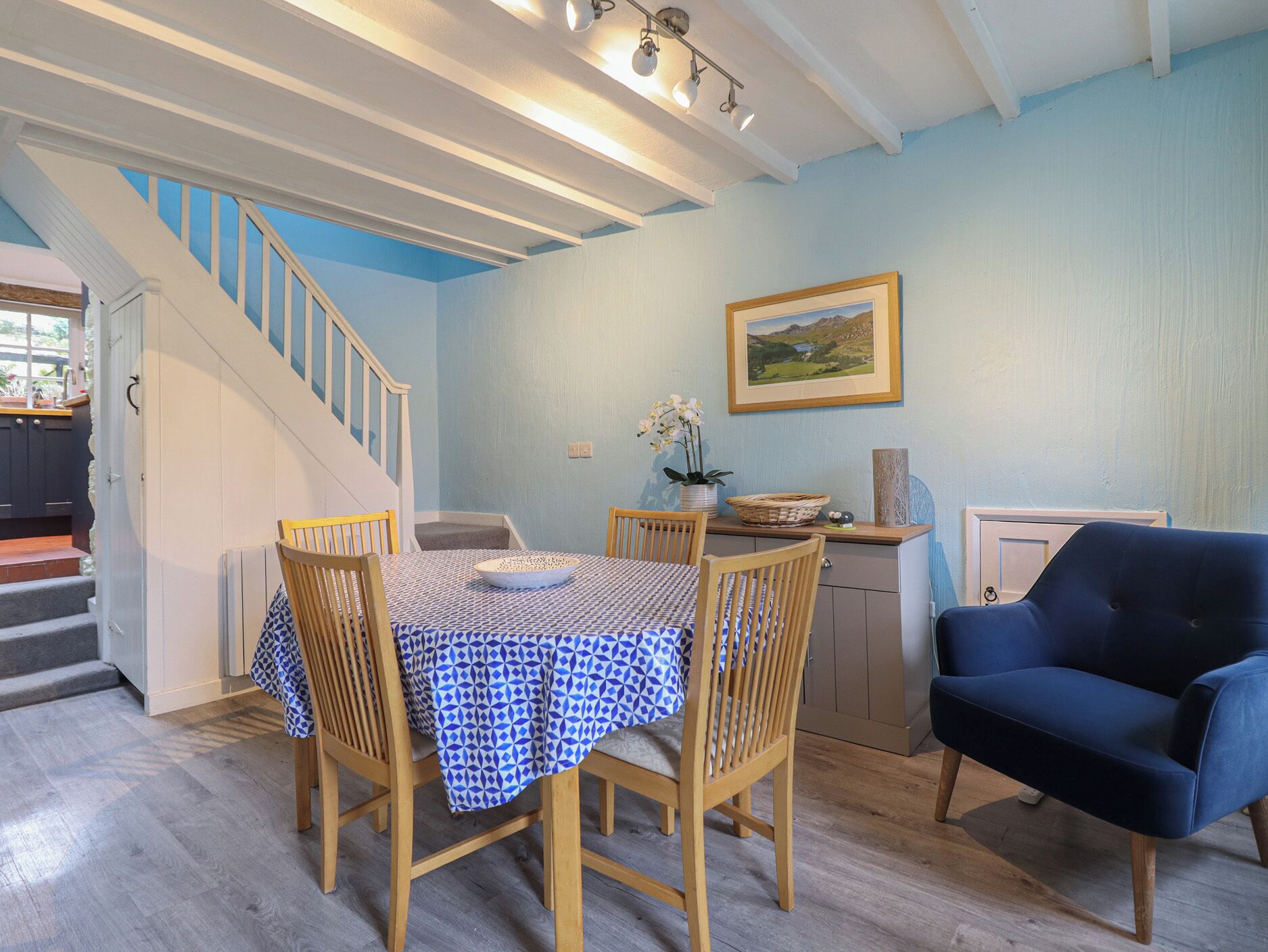 A dining area with a table set for four, wooden chairs, a blue and white patterned tablecloth, a blue sofa, a staircase, and a framed landscape painting on the wall.