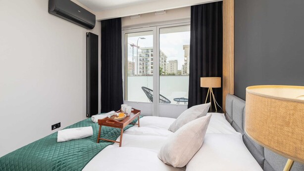 A hotel room with a bed, a window with curtains, a view of buildings, a table with a tray of breakfast, and a lamp.