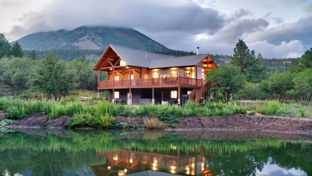 A wooden lodge with a balcony overlooking a calm lake, surrounded by lush greenery and mountains in the background. - Bears Ears Luxury Retreat (Monticello)