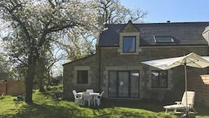 Une maison en pierre avec une grande fenêtre, une terrasse avec une table et des chaises, et un arbre dans la cour.