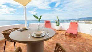 Een terras met een tafel voor twee personen, twee stoelen, een bloempot en uitzicht op zee.