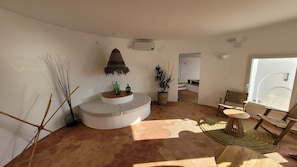 Ein Wohnzimmer mit einem runden weißen Sofa, einem kleinen Holztisch, einer Topfpflanze und einem Korbstuhl.