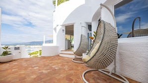 A terrace with hexagonal tiles, white walls, and a view of the city. There are two hanging chairs with patterned cushions and a hose on the floor. - Agi Castell Mar 1009 Apartamento for rent in Roses Girona. (Roses)