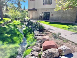 Zona ajardinada con un pequeño arroyo, rocas y un edificio con puerta y ventanas.