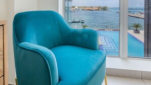 A teal armchair in a room with a large window overlooking a marina and a body of water. - NEW Modern Oceanfront Luxury Complex in Aruba! (Oranjestad)