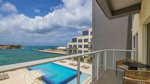 A balcony with a pool, wicker furniture, and a view of the ocean and buildings. - NEW Modern Oceanfront Luxury Complex in Aruba! (Oranjestad)