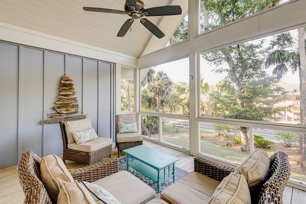 Relax in this serene sunroom with a high ceiling, ceiling fan, and expansive windows offering views of palm trees and a nearby building.