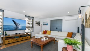 Una sala de estar moderna con un televisor de pantalla plana grande, un sofá blanco, una mesa de centro de madera y una planta en maceta.