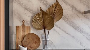 Una cocina moderna con protector contra salpicaduras de mármol, tabla de cortar de madera, un cuenco y un florero de vidrio con hojas secas.
