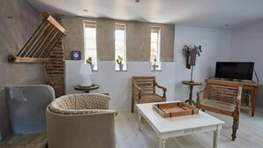 Una sala de estar con un sofá, dos sillas de madera, una mesa blanca, un televisor y una mesita de noche con una lámpara.