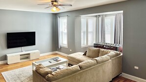 A living room with a sectional sofa, a flat-screen TV, a ceiling fan, and large windows with blinds.